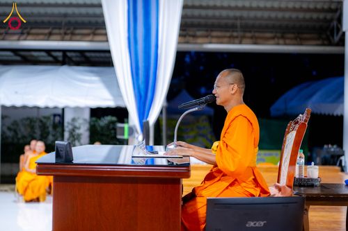 พระภิกษุสวมจีวรสีส้มกำลังนั่งบรรยายธรรมที่โต๊ะไม้หน้าไมโครโฟนภายในสถานที่จัดกิจกรรม