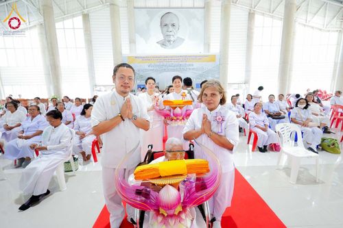 กลุ่มคนในชุดขาวร่วมพิธีทางศาสนาภายในอาคาร โดยมีผู้ถือพานผ้าไตรและพนมมือแสดงความเคารพ