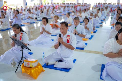 กลุ่มคนในชุดสีขาวนั่งพนมมือปฏิบัติธรรมโดยมีผู้นำสวดมนต์อยู่ด้านหน้า