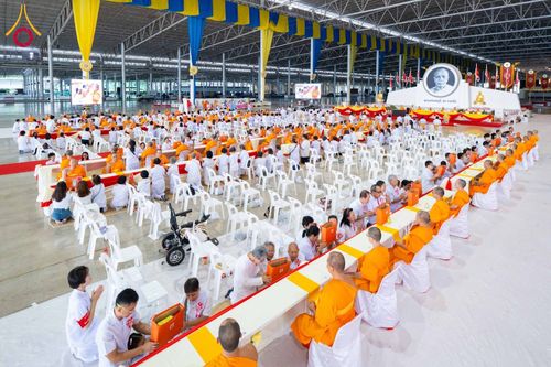 พุทธศาสนิกชนจำนวนมากนั่งเก้าอี้สีขาวร่วมพิธีถวายสังฆทานและทอดผ้าป่าแด่พระภิกษุสงฆ์ภายในอาคารขนาดใหญ่