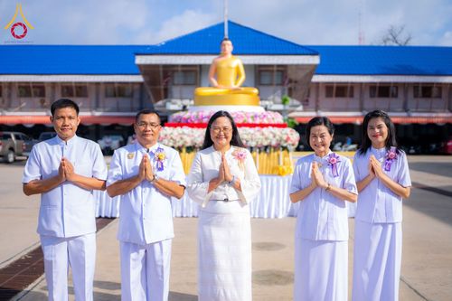 กลุ่มบุคคลชายหญิงสวมชุดสีขาวพนมมือไหว้ ด้านหลังเป็นพระพุทธรูปและอาคาร