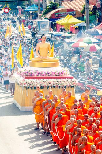 ขบวนแห่พระพุทธรูปสีทองบนรถบุษบกประดับดอกไม้ มีพระภิกษุสงฆ์เดินนำขบวนและพุทธศาสนิกชนร่วมเดินตามบนถนน