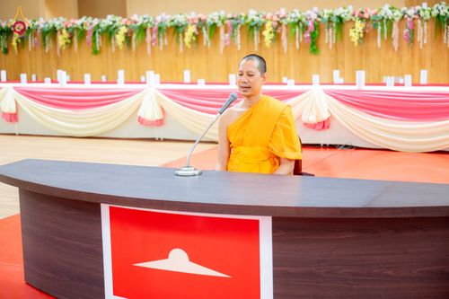 พระภิกษุสงฆ์นั่งบรรยายธรรมที่โต๊ะไม้หน้าไมโครโฟน ภายในห้องโถงที่มีการตกแต่งด้วยผ้าและดอกไม้