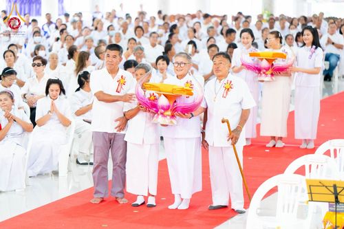 กลุ่มพุทธศาสนิกชนในชุดขาวถือพานพุ่มผ้าไตรยืนบนพรมแดงในพิธีทางศาสนา
