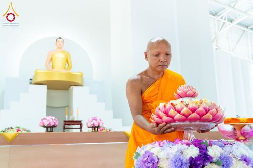 พระภิกษุสงฆ์ถือพานดอกไม้สีชมพูในพิธีทางศาสนา โดยมีพระพุทธรูปสีทองเป็นฉากหลัง