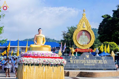 พระพุทธรูปสีทองประดิษฐานบนฐานดอกไม้สด ตั้งอยู่หน้าป้ายที่ว่าการอำเภอท่าตะโกและซุ้มตราสัญลักษณ์