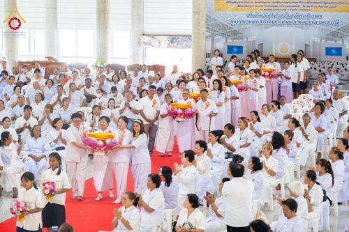 กลุ่มพุทธศาสนิกชนสวมชุดขาวร่วมพิธีทางศาสนาภายในอาคาร มีผู้ถือพานพุ่มดอกบัวเดินนำขบวนท่ามกลางผู้เข้าร่วมพิธีที่นั่งพนมมือ