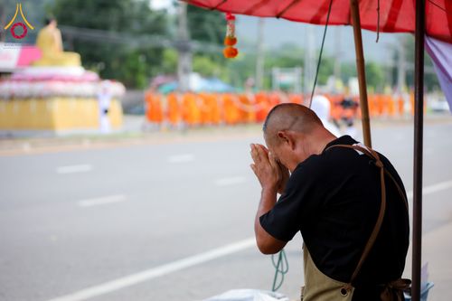 ชายไทยพนมมือไหว้หันข้าง โดยมีขบวนพระภิกษุสงฆ์เดินอยู่บนถนนเป็นฉากหลัง