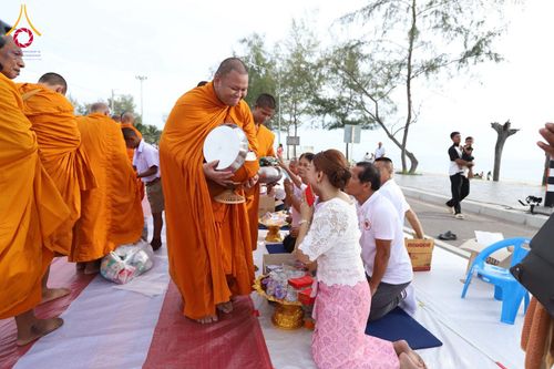 พระสงฆ์ในจีวรสีส้มกำลังรับบิณฑบาตจากพุทธศาสนิกชนที่นั่งคุกเข่าอยู่บนพื้นริมชายหาด