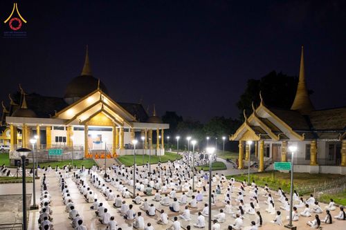 ผู้คนสวมชุดขาวนั่งสมาธิบนลานกว้างหน้าอาคารทรงไทยในยามค่ำคืน