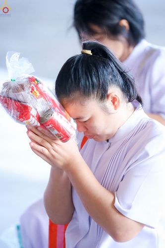 หญิงสาวสวมชุดสีขาวกำลังพนมมือถือถุงใส่ของตักบาตรด้วยท่าทางสงบและตั้งใจ
