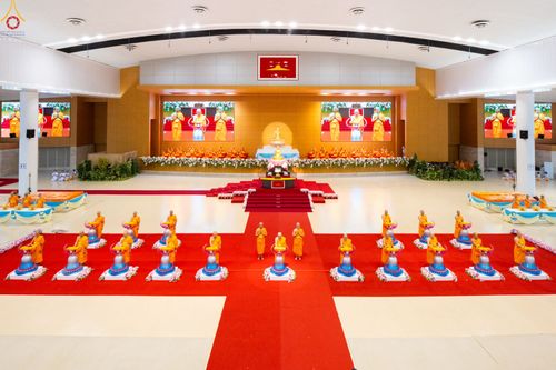 พระภิกษุสงฆ์ยืนประกอบพิธีทอดผ้าป่าสมทบกฐินภายในสภาธรรมกายสากลที่มีพรมสีแดงและฉากหลังเป็นจอภาพขนาดใหญ่