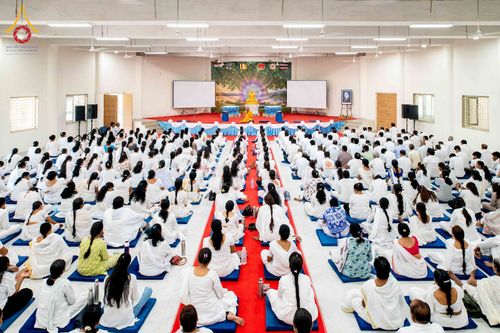 ผู้คนจำนวนมากสวมชุดสีขาวนั่งปฏิบัติธรรมในหอประชุมขนาดใหญ่โดยมีพระภิกษุสงฆ์อยู่บนเวที
