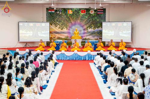พระภิกษุสงฆ์และพุทธศาสนิกชนชุดขาวกำลังร่วมกิจกรรมปฏิบัติธรรมภายในหอประชุมที่มีฉากหลังเป็นภาพพระพุทธรูป