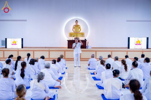 วิทยากรชายสวมชุดขาวกำลังบรรยายธรรมะต่อหน้าผู้เข้าอบรมชุดขาวจำนวนมากในห้องโถง