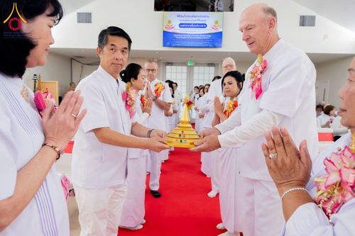 กลุ่มคนในชุดขาวกำลังถือผอบทองคำจำลองในพิธีทางศาสนาภายในอาคารที่มีพรมแดง