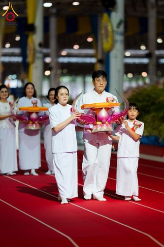 กลุ่มคนสวมชุดขาวถือพานพุ่มผ้าไตรเดินบนพรมสีแดงในพิธีทางศาสนา