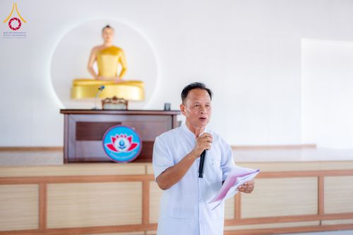 ชายสวมชุดขาวถือไมโครโฟนและเอกสารกำลังพูดบรรยายหน้าพระพุทธรูป