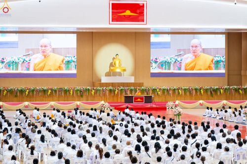 ผู้เข้าร่วมงานสวมชุดขาวนั่งฟังธรรมภายในหอประชุมที่มีพระภิกษุสงฆ์แสดงธรรมบนเวทีและมีจอภาพขนาดใหญ่