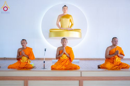 พระภิกษุสงฆ์ 3 รูปนั่งพนมมือปฏิบัติธรรมบนเวทีไม้เบื้องหน้าพระพุทธรูปสีทอง