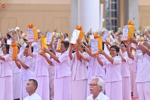กลุ่มพุทธศาสนิกชนสวมชุดขาวกำลังชูห่อของขวัญสีทองเหนือศีรษะในพิธีทางศาสนา
