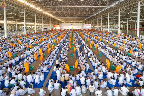 ผู้คนจำนวนมากสวมชุดขาวนั่งเรียงแถวตักบาตรพระสงฆ์ภายในอาคารกว้างขวาง