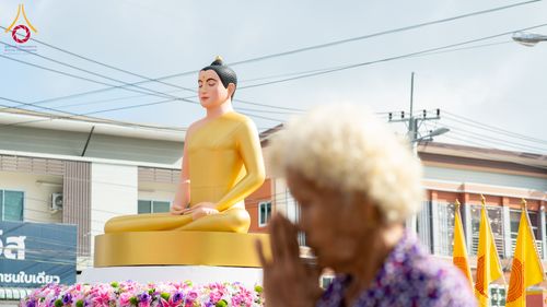พระพุทธรูปปางสมาธิสีทองประดิษฐานบนฐานดอกไม้ โดยมีหญิงสูงวัยยืนพนมมือไหว้ในมุมมองเบลออยู่ด้านหน้า