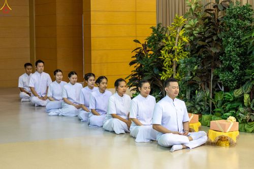 กลุ่มคนสวมชุดสีขาวนั่งสมาธิเรียงแถวกันบนพื้นในอาคาร
