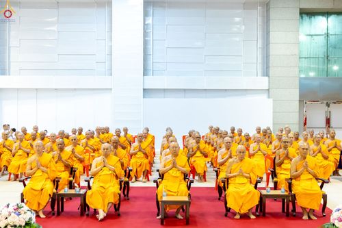 คณะสงฆ์จำนวนมากในจีวรสีเหลืองทองนั่งพนมมือบนเก้าอี้ภายในอาคารสภาธรรมกายสากล