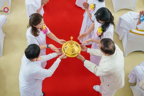 กลุ่มคนในชุดขาวกำลังช่วยกันถือพานพุ่มสีทองประดับอัญมณีเหนือพรมสีแดง