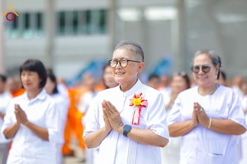 ผู้หญิงสวมชุดสีขาวพนมมือไหว้ด้วยใบหน้ายิ้มแย้มท่ามกลางผู้เข้าร่วมพิธีคนอื่นๆ