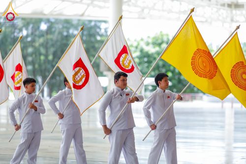 กลุ่มชายหนุ่มสวมชุดสีขาวถือธงสัญลักษณ์สีขาวและสีเหลืองเดินเรียงแถวในอาคาร