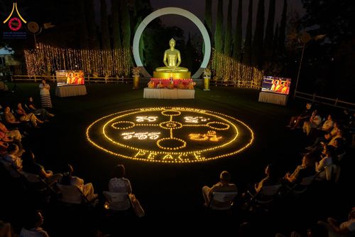 ผู้คนนั่งล้อมวงรอบการจุดประทีปเป็นรูปสัญลักษณ์และคำว่า PEACE หน้าพระพุทธรูปสีทองในยามค่ำคืน