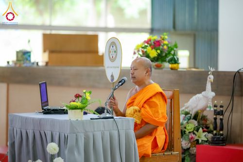 พระภิกษุสงฆ์กำลังนั่งบรรยายธรรมหน้าโต๊ะที่มีคอมพิวเตอร์โน้ตบุ๊กและพัดยศ
