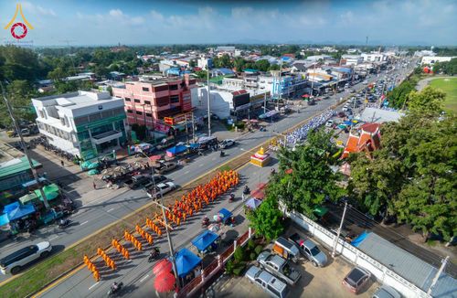 ขบวนพระภิกษุสงฆ์ห่มจีวรสีส้มเดินเรียงแถวบนถนนในเขตชุมชน โดยมีรถบุษบกอัญเชิญพระพุทธรูปนำหน้าขบวน