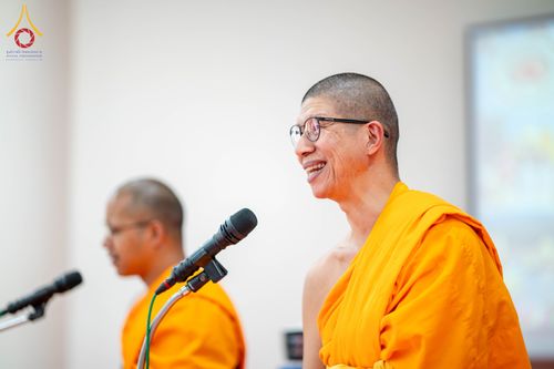 พระภิกษุสงฆ์สวมแว่นตากำลังพูดผ่านไมโครโฟนด้วยท่าทางยิ้มแย้ม