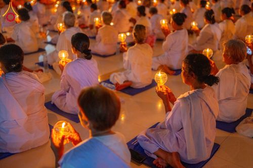 กลุ่มอุบาสิกาในชุดขาวนั่งสมาธิถือประทีปในมือภายในอาคาร