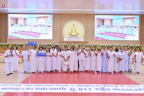 กลุ่มคนสวมชุดสีขาวจำนวนมากยืนเรียงแถวบนเวทีในห้องประชุมขนาดใหญ่