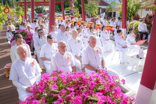 กลุ่มคนสวมชุดขาวนั่งร่วมพิธีทางศาสนาพุทธในพื้นที่กลางแจ้งที่มีดอกไม้สีชมพูประดับอยู่ด้านหน้า