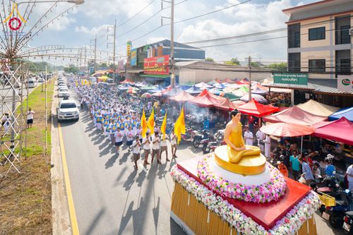 ขบวนแห่พระพุทธรูปสีทองบนรถบุษบกและกลุ่มคนสวมชุดสีขาวเดินบนถนนที่มีร้านค้า