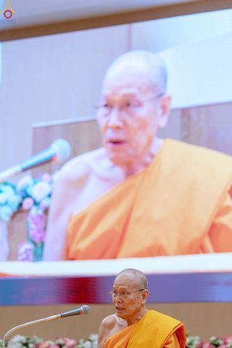 พระภิกษุสงฆ์กำลังเทศนาธรรมโดยมีภาพของท่านบนจอโปรเจกเตอร์ด้านหลัง