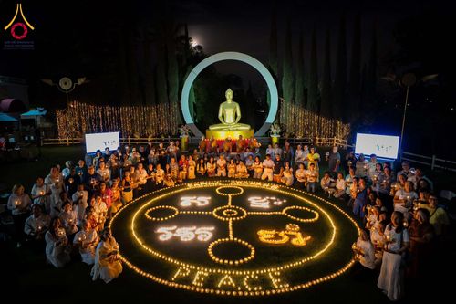 กลุ่มคนนั่งล้อมรอบวงกลมแสงเทียนและคำว่า PEACE หน้าพระพุทธรูปสีทองในยามค่ำคืน