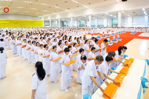 กลุ่มคนจำนวนมากสวมชุดสีขาวกำลังเข้าแถวเพื่อวางผ้าไตรในพิธีทางศาสนาภายในอาคารขนาดใหญ่