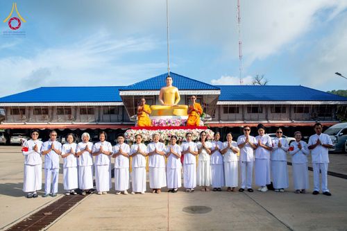 กลุ่มคนชุดขาวและพระภิกษุสงฆ์ยืนหน้าองค์พระประธานสีทองในพิธีทางศาสนา