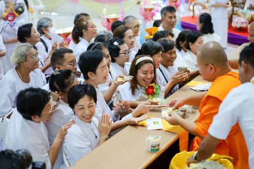 พระภิกษุสงฆ์ในจีวรสีส้มกำลังมอบวัตถุมงคลให้แก่กลุ่มพุทธศาสนิกชนที่สวมชุดขาวภายในอาคาร