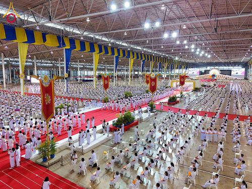 พุทธศาสนิกชนสวมชุดขาวจำนวนมากนั่งร่วมพิธีในอาคารขนาดใหญ่ที่มีการตกแต่งด้วยธงและพรมสีแดง