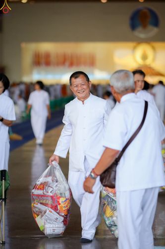 ชายไทยสวมชุดขาวถือถุงใส่ข้าวสารอาหารแห้งเดินยิ้มแย้มภายในอาคาร