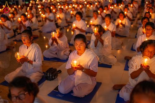 กลุ่มอุบาสิกาสวมชุดขาวนั่งสมาธิถือประทีปส่องสว่างในมือท่ามกลางบรรยากาศที่สงบ