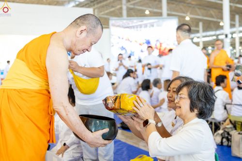 หญิงสูงวัยสวมชุดสีขาวกำลังประเคนอาหารแห้งใส่บาตรพระภิกษุสงฆ์ในพิธีตักบาตร