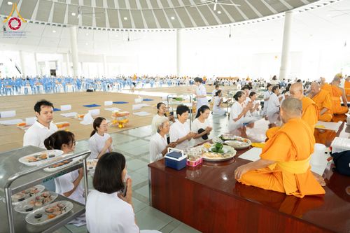 กลุ่มคนในชุดขาวกำลังประนมมือถวายภัตตาหารแด่พระภิกษุสงฆ์ภายในอาคารโถงกว้าง
